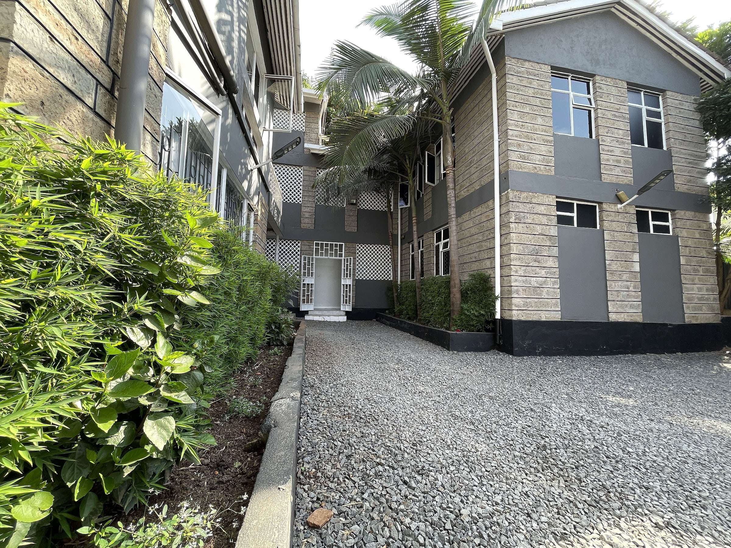 A gravel path between leafy hedges and palms, leading into the Maskan compound